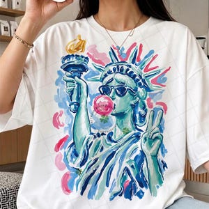 May include: White t-shirt featuring a colorful illustration of the Statue of Liberty wearing sunglasses and blowing a bubblegum bubble. The design includes blue, pink, and yellow accents, with the text "ALL RIGHTS PROTECTED COPYRIGHT" visible.