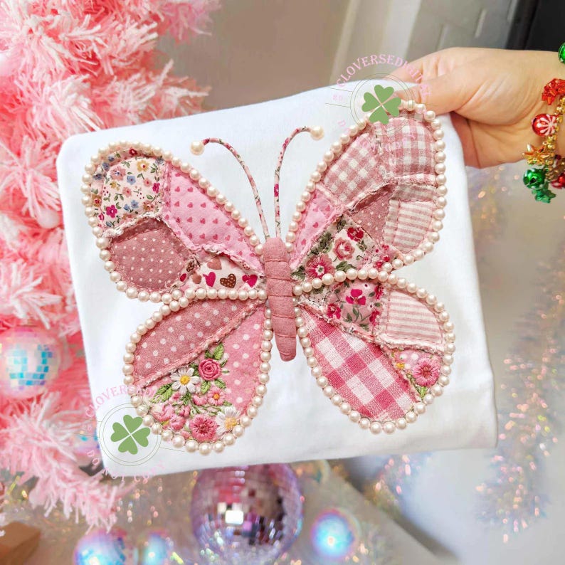 May include: A white t-shirt featuring a large, pink butterfly applique. The butterfly has patchwork wings with floral, polka dot, and gingham patterns, outlined with faux pearls. The butterfly's body is a solid pink, and the antennae have pearl accents.