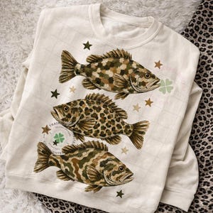 May include: Cream-colored sweatshirt featuring three detailed fish illustrations in shades of brown, green, and tan. The fish have unique patterns, with one displaying a leopard-like design. Scattered around the fish are brown star shapes. The sweatshirt has a crew neck.