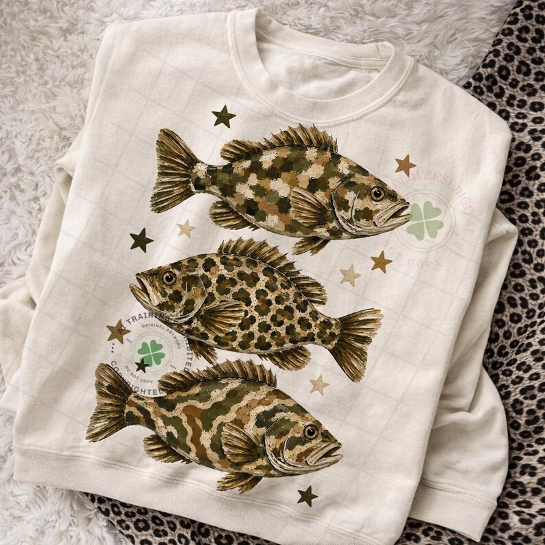 May include: Cream-colored sweatshirt featuring three detailed fish illustrations in shades of brown, green, and tan. The fish have unique patterns, with one displaying a leopard-like design. Scattered around the fish are brown star shapes. The sweatshirt has a crew neck.