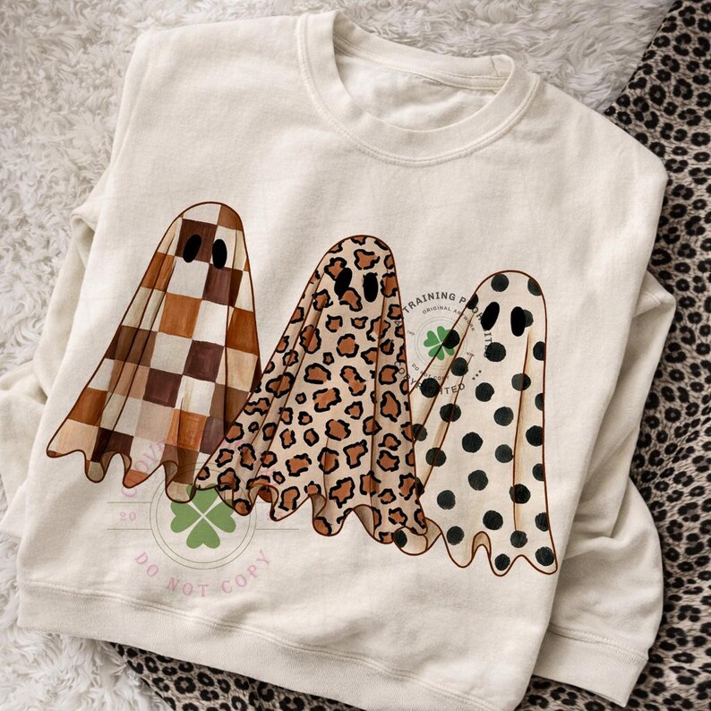 May include: A cream-colored crewneck sweatshirt featuring a graphic of three ghosts. The first ghost has a brown and white checkered pattern, the second has a brown leopard print, and the third has black polka dots on a cream background.
