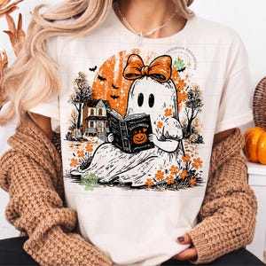 Op de afbeelding: Een cr&egrave;mekleurig t-shirt met een afbeelding van een geest met een oranje strik, die een boek leest met de titel Halloween en een pompoenillustratie. De sc&egrave;ne bevat een huis, bomen, vleermuizen en oranje bloemen tegen een oranje zon.