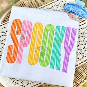 以下が含まれることがあります： 白のTシャツに、大きくカラフルなストライプ柄の文字で「SPOOKY」とプリントされています。各文字はオレンジ、ピンク、イエロー、グリーン、ティール、パープルの異なる色で、ジグザグのステッチ縁取りが施されています。水色のヘアクリップが一緒に写っています。