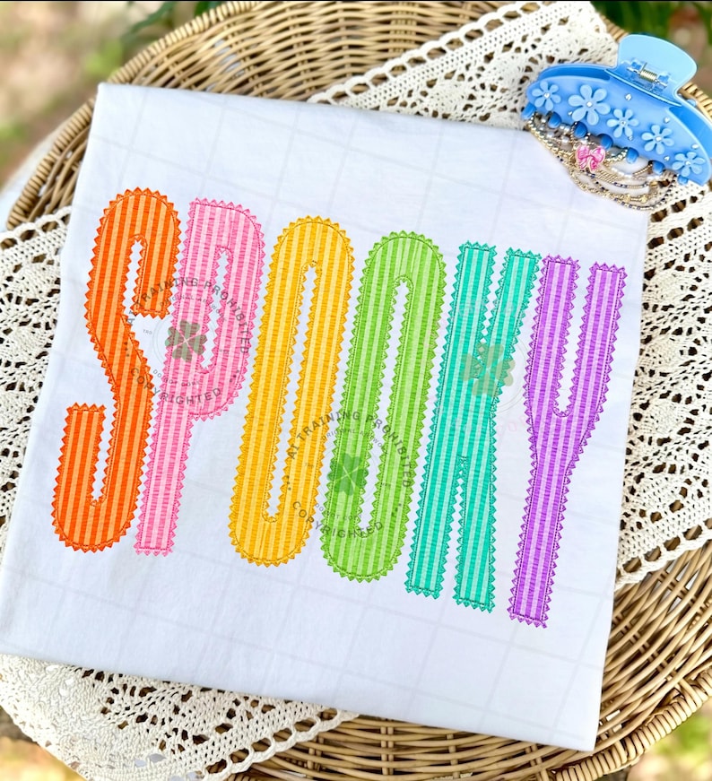 以下が含まれることがあります： 白のTシャツに、大きくカラフルなストライプ柄の文字で「SPOOKY」とプリントされています。各文字はオレンジ、ピンク、イエロー、グリーン、ティール、パープルの異なる色で、ジグザグのステッチ縁取りが施されています。水色のヘアクリップが一緒に写っています。