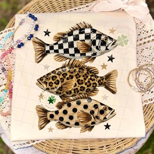 May include: A cream-colored t-shirt featuring three stylized fish with unique patterns: a black and white checkerboard, leopard print, and black polka dots. The design includes stars and a four-leaf clover.