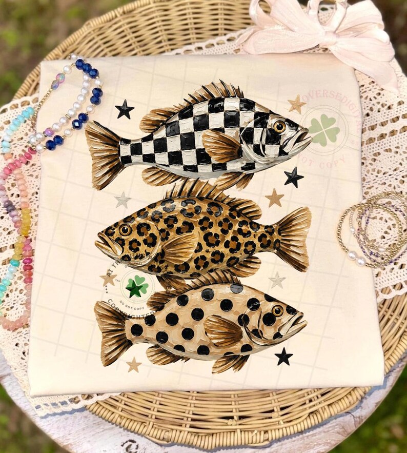May include: A cream-colored t-shirt featuring three stylized fish with unique patterns: a black and white checkerboard, leopard print, and black polka dots. The design includes stars and a four-leaf clover.