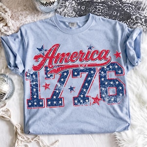 Może przedstawiać: Jasnoniebieska koszulka z napisem "America" w czerwonym skrypcie i liczbami "1776" w kolorze niebieskim z białymi gwiazdami. Koszulka ma wygląd vintage, idealna na patriotyczne wydarzenia.