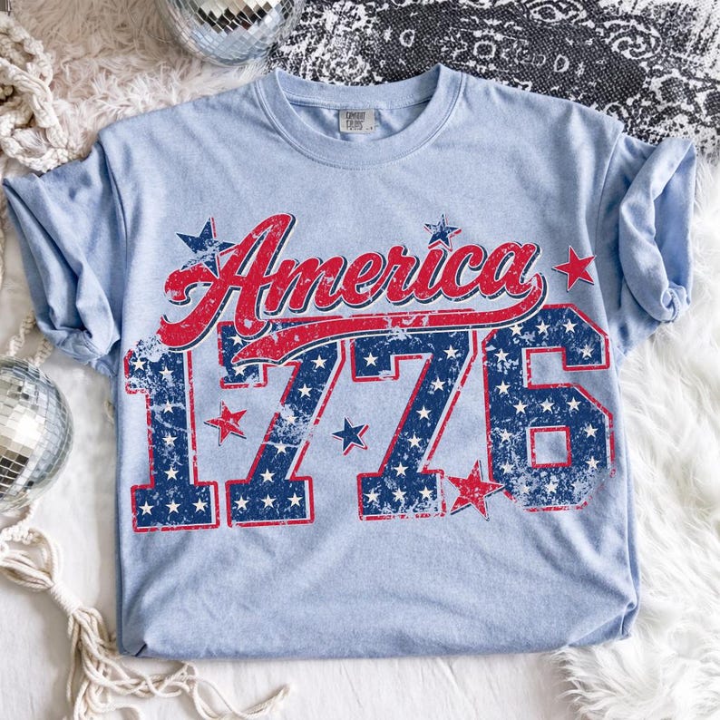 Może przedstawiać: Jasnoniebieska koszulka z napisem "America" w czerwonym skrypcie i liczbami "1776" w kolorze niebieskim z białymi gwiazdami. Koszulka ma wygląd vintage, idealna na patriotyczne wydarzenia.