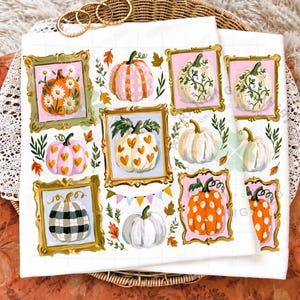 May include: A white fabric surface featuring a repeating pattern of various pumpkins inside ornate gold frames. The pumpkins display diverse designs including daisies, pink and white plaid, gold hearts, black and white buffalo check, and orange polka dots.