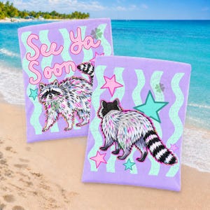 May include: Two lavender beach towels with a raccoon illustration and the text "See Ya Soon". The towels feature a wavy pattern in light blue and are decorated with stars and four-leaf clovers. The towels are on a sandy beach.