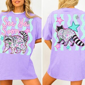May include: A lilac-colored t-shirt featuring a raccoon graphic with the text "See Ya Soon". The raccoon is depicted with colorful fur and a striped tail. The back of the shirt also has a raccoon graphic and stars.