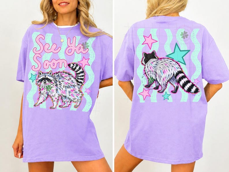 May include: A lilac-colored t-shirt featuring a raccoon graphic with the text "See Ya Soon". The raccoon is depicted with colorful fur and a striped tail. The back of the shirt also has a raccoon graphic and stars.