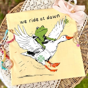 May include: A light yellow t-shirt featuring a whimsical illustration of a green frog riding a white goose with orange feet and beak. The text "we ride at dawn" is above the illustration. The shirt is displayed on a wicker basket.