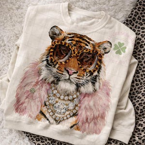 May include: Cream-colored sweatshirt featuring a tiger wearing leopard print sunglasses, a pink feather boa, and a diamond necklace. The tiger's face is detailed with orange and black stripes. The design is a whimsical and fashionable statement.
