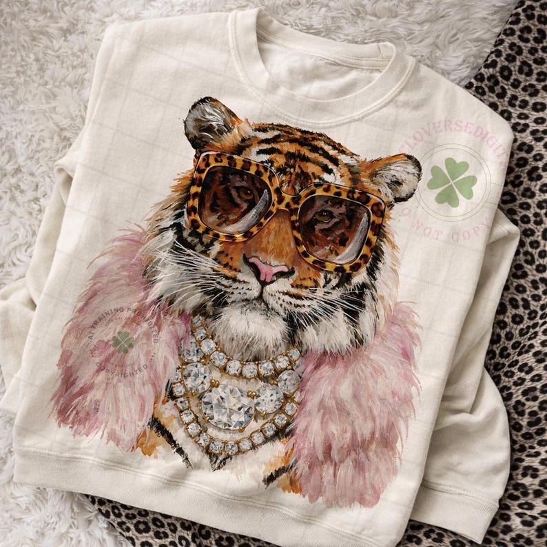 May include: Cream-colored sweatshirt featuring a tiger wearing leopard print sunglasses, a pink feather boa, and a diamond necklace. The tiger's face is detailed with orange and black stripes. The design is a whimsical and fashionable statement.