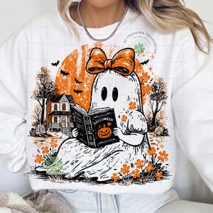 Op de afbeelding: Een wit sweatshirt met lange mouwen en een afbeelding van een geest met een oranje strik die een boek leest met de titel Halloween. Het ontwerp bevat een oranje maan, een huis, vleermuizen en oranje bloemen op de voorzijde van het kledingstuk.