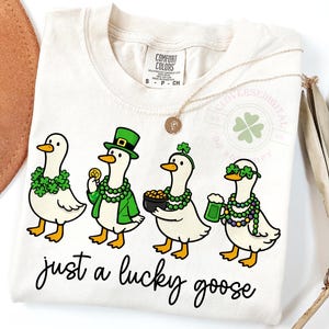 May include: A cream-coloured t-shirt with a St. Patrick's Day design. It shows four cartoon geese adorned with green shamrocks, hats, and beads. The phrase "just a lucky goose" is written in a script font below the geese.