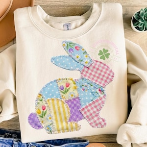 May include: A cream-coloured sweatshirt with a patchwork bunny design. The bunny is made from various patterned fabrics, including polka dots, floral prints, and gingham. The design incorporates pastel colours like blue, pink, yellow, and purple, creating a spring-themed aesthetic.