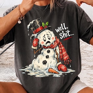 May include: A charcoal gray t-shirt featuring a graphic of a melting snowman wearing a black top hat with a red band and holly, a red plaid scarf, and red mittens. The snowman has a sad expression and the text well, shit... is printed in white.
