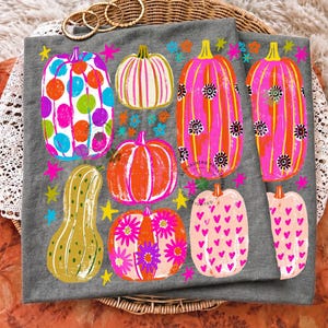 May include: A grey fabric item featuring a colourful graphic print of various pumpkins. The pumpkins are decorated with patterns including polka dots, stripes, flowers, and hearts in bright shades of pink, orange, blue, and yellow, accented by stars.