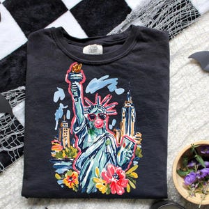 May include: Black crew neck t-shirt featuring a colorful illustration of the Statue of Liberty wearing sunglasses and blowing a bubble. The design includes the New York City skyline and floral accents. The shirt has the text "Conway Colors" on the tag.