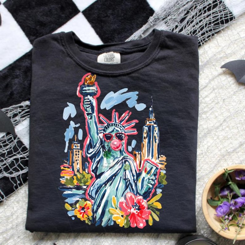 May include: Black crew neck t-shirt featuring a colorful illustration of the Statue of Liberty wearing sunglasses and blowing a bubble. The design includes the New York City skyline and floral accents. The shirt has the text "Conway Colors" on the tag.