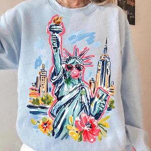 May include: Light blue sweatshirt featuring a colorful illustration of the Statue of Liberty wearing sunglasses and blowing a bubble. The design includes the New York City skyline and floral accents. The word "COPYRIGHT" is visible on the design.