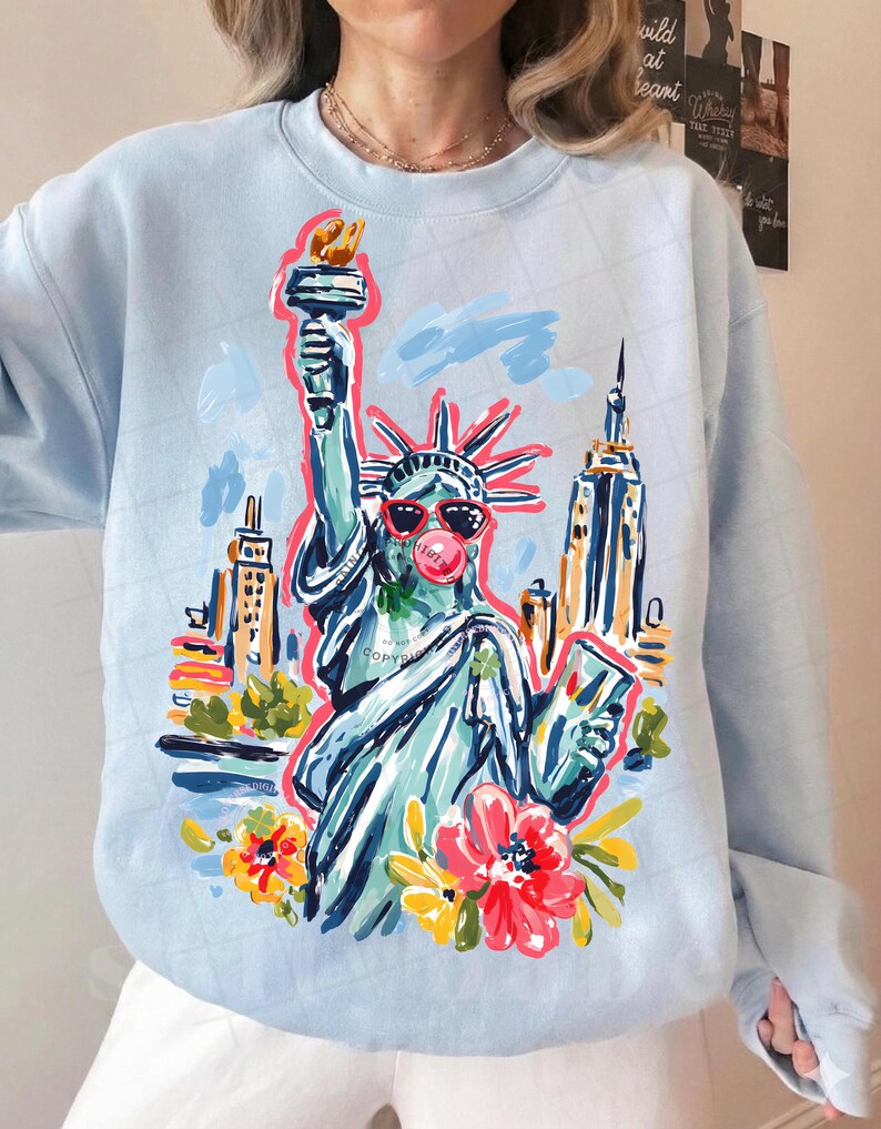 May include: Light blue sweatshirt featuring a colorful illustration of the Statue of Liberty wearing sunglasses and blowing a bubble. The design includes the New York City skyline and floral accents. The word "COPYRIGHT" is visible on the design.
