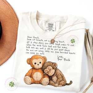 May include: Cream-colored Comfort Colors t-shirt featuring a graphic of two plush monkeys, one brown and one tan, with the text "Dear Punch, I am not breath..." and "Love, Your Plush". A gold necklace is draped across the shirt.