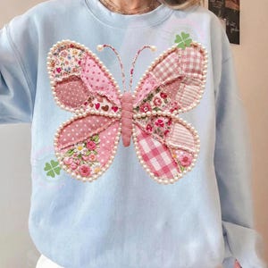 May include: Light blue sweatshirt featuring a large butterfly design. The butterfly is made of pink patchwork fabric with floral and polka dot patterns, outlined with faux pearls. The butterfly's body is a textured pink fabric.