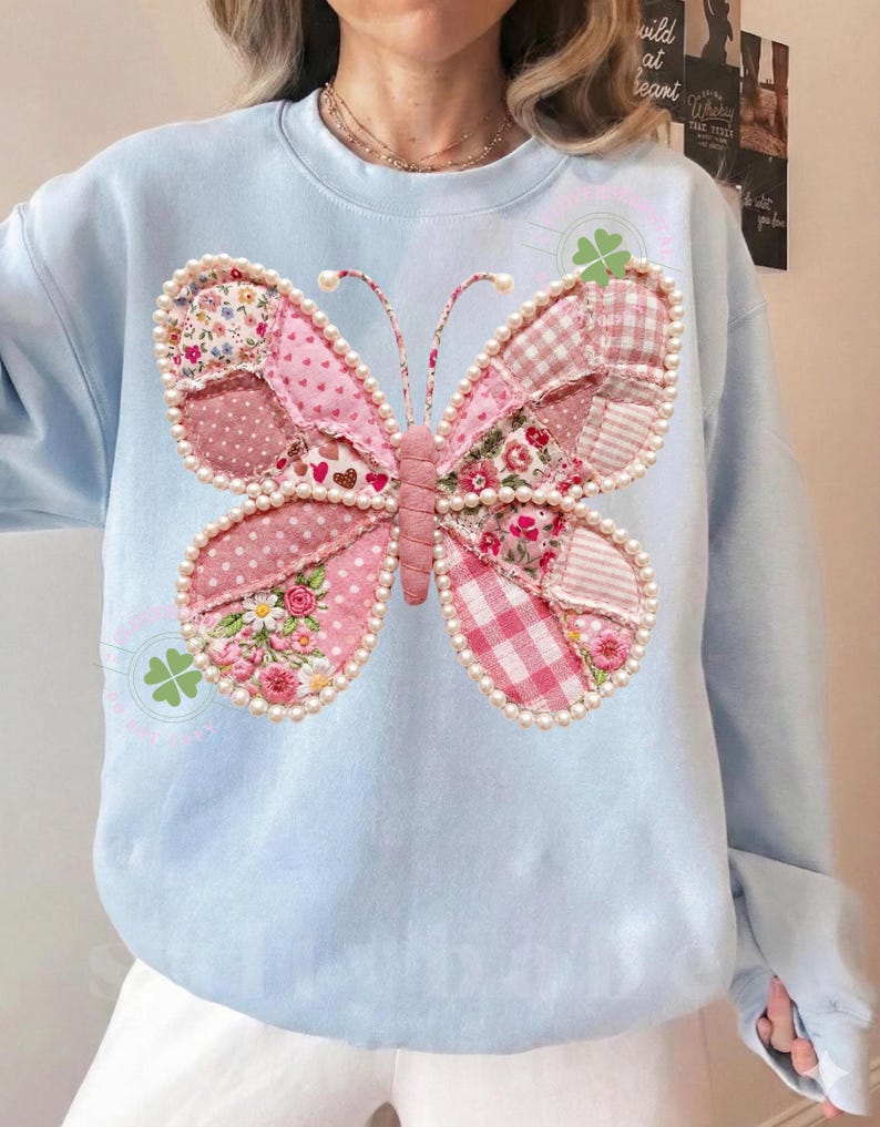 May include: Light blue sweatshirt featuring a large butterfly design. The butterfly is made of pink patchwork fabric with floral and polka dot patterns, outlined with faux pearls. The butterfly's body is a textured pink fabric.