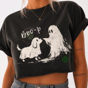 May include: A charcoal black short-sleeve t-shirt with a raw hem. The front features a white graphic of a ghost and a dog wearing a ghost costume, with the text "BOO" printed above them. The design includes small green clover icons.