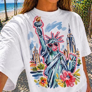 May include: White t-shirt featuring a colorful illustration of the Statue of Liberty wearing sunglasses and blowing a bubblegum bubble. The design includes the New York City skyline and flowers.