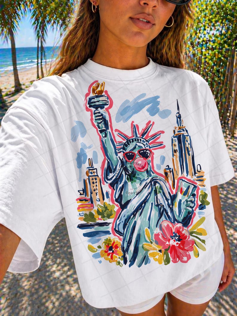 May include: White t-shirt featuring a colorful illustration of the Statue of Liberty wearing sunglasses and blowing a bubblegum bubble. The design includes the New York City skyline and flowers.