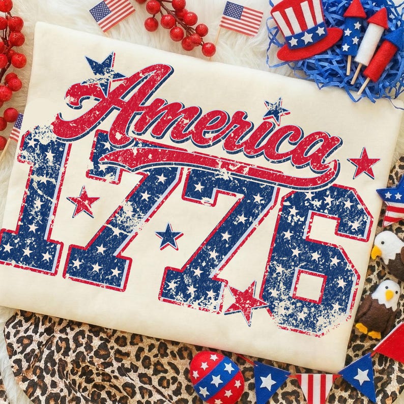 Może przedstawiać: Kremowa koszulka z postarzaną, czerwono-niebieską grafiką z napisem "America 1776". Projekt zawiera gwiazdy i paski, nawiązując do patriotycznego motywu. Dodatkowe patriotyczne dekoracje obejmują miniaturowe flagi i świąteczne ozdoby.