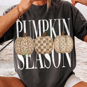 May include: A charcoal gray short-sleeve t-shirt featuring the white text PUMPKIN SEASON in a distressed font. Between the words are three pumpkins with different patterns: one with a leopard print, one with a checkered pattern, and one with polka dots.