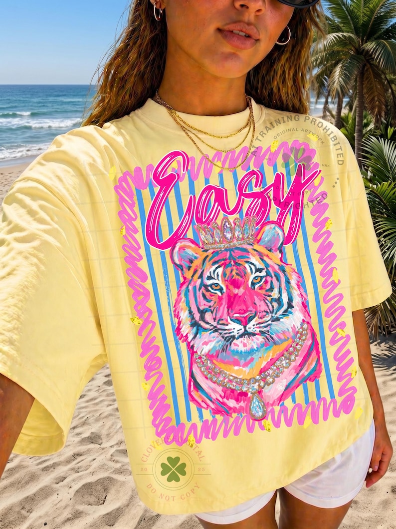 May include: Yellow t-shirt with a colorful tiger graphic wearing a crown and necklace. The word "Easy" is written in pink script above the tiger. The design is framed by blue stripes and pink squiggles.