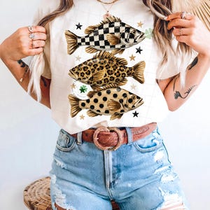 May include: Cream-colored t-shirt featuring three fish designs with unique patterns: checkered, leopard print, and polka dots. Each fish is accompanied by star and shamrock accents. The shirt is paired with denim shorts and a brown belt.
