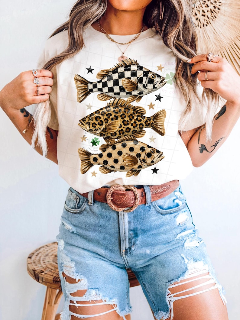 May include: Cream-colored t-shirt featuring three fish designs with unique patterns: checkered, leopard print, and polka dots. Each fish is accompanied by star and shamrock accents. The shirt is paired with denim shorts and a brown belt.