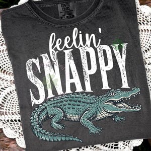 May include: A charcoal grey short-sleeve t-shirt featuring the white text "feelin' SNAPPY" in a distressed font. Below the text is a detailed illustration of a green alligator with its mouth open, showing sharp teeth and textured scales.