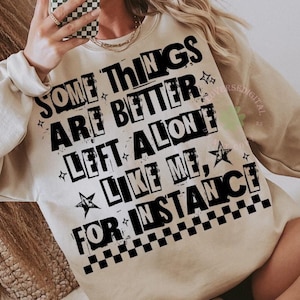 Puede incluir: Sudadera beige con el texto negro "SOME THINGS ARE BETTER LEFT ALONE LIKE ME, FOR INSTANCE". El diseño incluye estrellas y cuadros. Una prenda superior informal y cómoda.