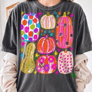 May include: A charcoal grey t-shirt featuring a graphic print of six colourful pumpkins in various patterns, including polka dots, stripes, flowers, and hearts, interspersed with small bright stars and flower shapes in neon colours.