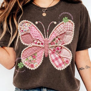May include: Dark brown t-shirt featuring a large, patchwork butterfly design. The butterfly has pink wings with various patterns, including polka dots, floral prints, and gingham. The design is outlined with faux pearls. Text on the shirt reads "CLOVERSEDIGITAL" and "NOT COPY".