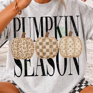 May include: A light gray crewneck sweatshirt featuring the black text PUMPKIN SEASON. Three pumpkins are centered in the design, each with a different pattern: one with a leopard print, one with a checkered pattern, and one with a polka dot pattern.