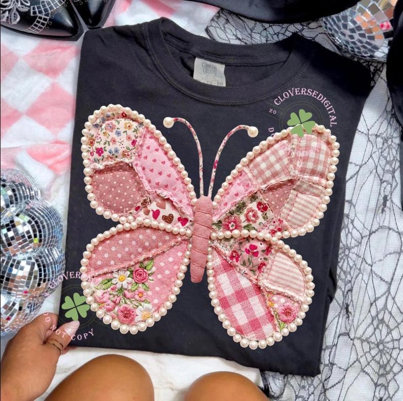 May include: Black t-shirt featuring a large, appliqued butterfly design. The butterfly has pink and floral patterned wings, outlined with pearls. The shirt includes the text "CLOVERSEDIGITAL" and a four-leaf clover logo.