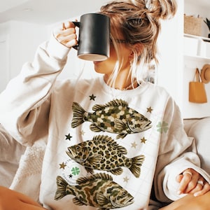May include: Cream-colored sweatshirt featuring three detailed fish illustrations in shades of brown and green, with star accents. The design includes a copyright symbol and a four-leaf clover. A black mug is held up.