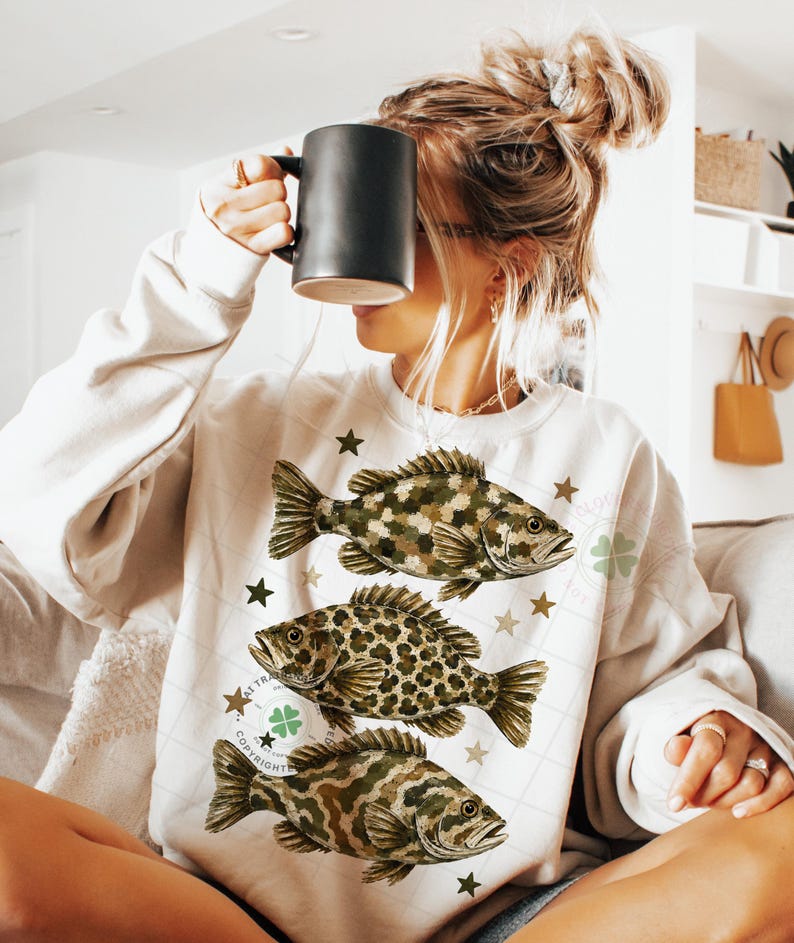 May include: Cream-colored sweatshirt featuring three detailed fish illustrations in shades of brown and green, with star accents. The design includes a copyright symbol and a four-leaf clover. A black mug is held up.