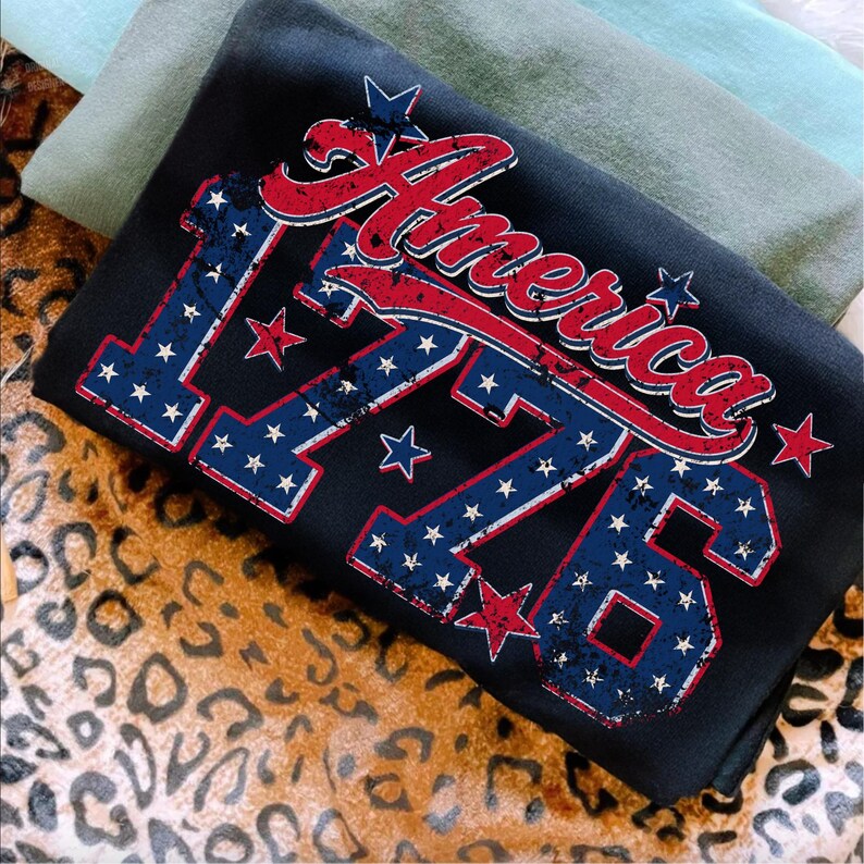 Może przedstawiać: Czarny t-shirt z napisem "America" w czerwonym skrypcie i numerem "1776" w kolorze niebieskim z białymi gwiazdami. Projekt ma postarzany, vintage wygląd. Koszulka jest złożona z innymi koszulkami w tle.