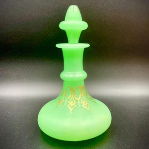 Pode incluir: Um frasco de perfume de vidro verde com uma base bulbosa e uma rolha decorativa. O frasco tem desenhos dourados ornamentados perto da base. O frasco tem aproximadamente 15 cm de altura.