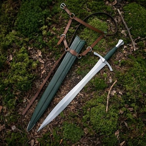 Puede incluir: Una espada de acero con empuñadura verde oscuro y una vaina verde oscuro a juego. La espada presenta una guarda y un pomo plateados. Un arnés de cuero marrón con hebillas y anillos de metal plateado está unido a la vaina para su transporte.